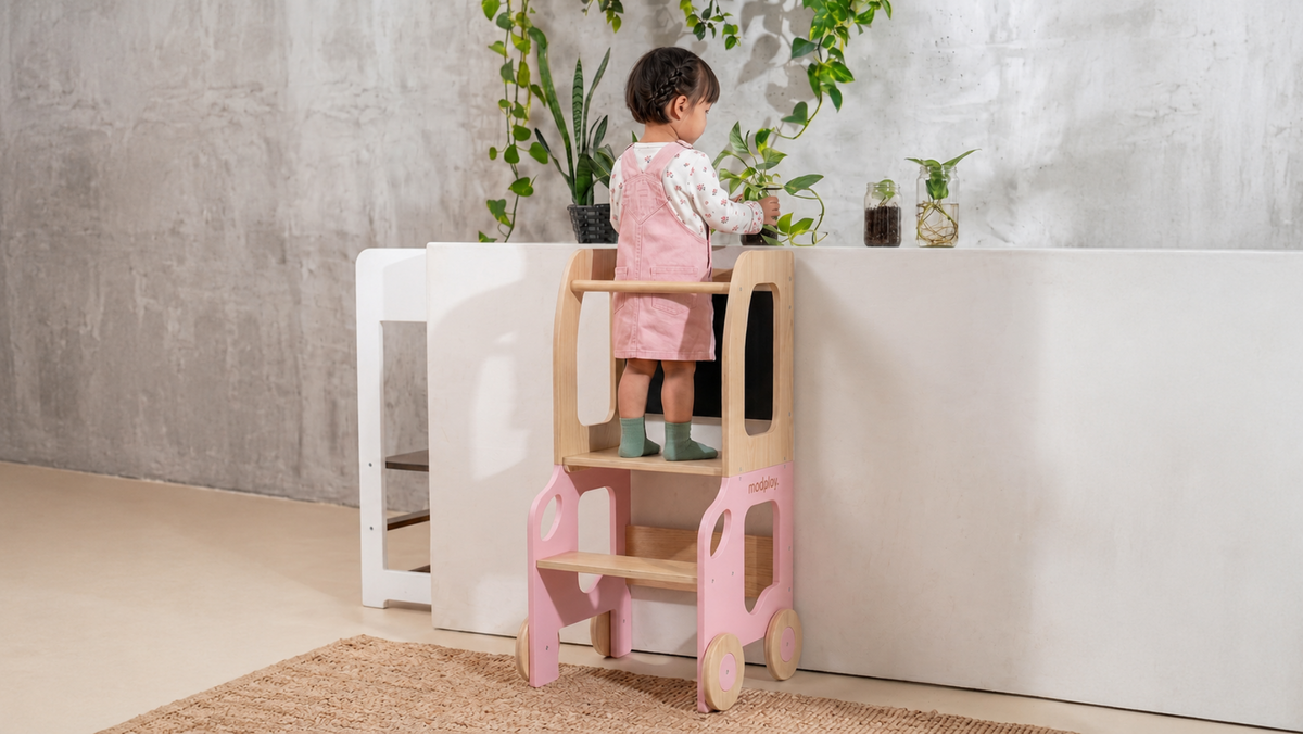 A toddler stands on a pink and natural wood step stool in a bright, modern room with a concrete-textured wall, white counter, and potted plants.
